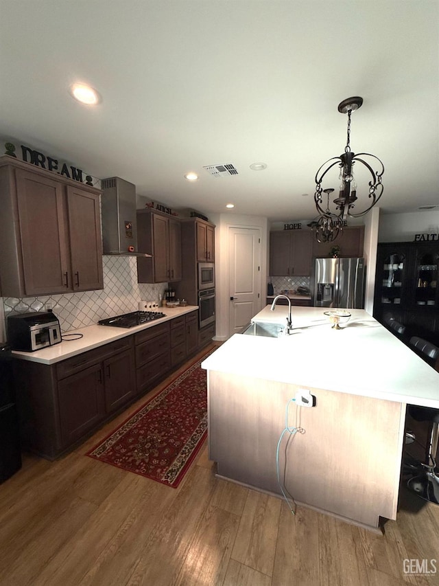 kitchen featuring wall chimney exhaust hood, an island with sink, light hardwood / wood-style floors, and appliances with stainless steel finishes