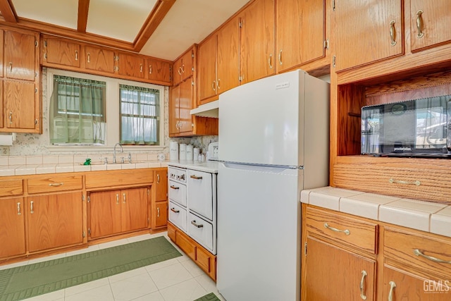 kitchen featuring backsplash, under cabinet range hood, light tile patterned floors, freestanding refrigerator, and a sink