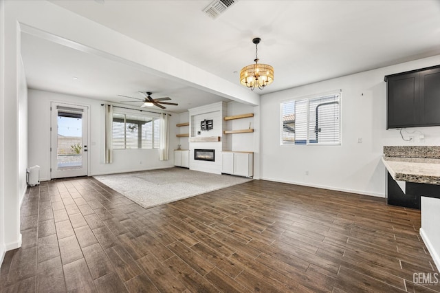 unfurnished living room featuring dark wood-style flooring, visible vents, baseboards, a fireplace, and ceiling fan with notable chandelier