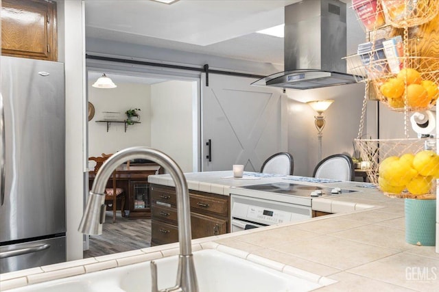 kitchen featuring tile countertops, stainless steel refrigerator, island exhaust hood, wall oven, and a barn door