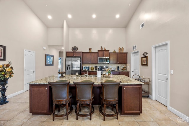 kitchen with appliances with stainless steel finishes, high vaulted ceiling, a large island with sink, light tile patterned floors, and dark brown cabinetry