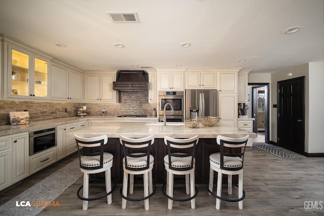 kitchen with backsplash, a center island with sink, custom range hood, and appliances with stainless steel finishes