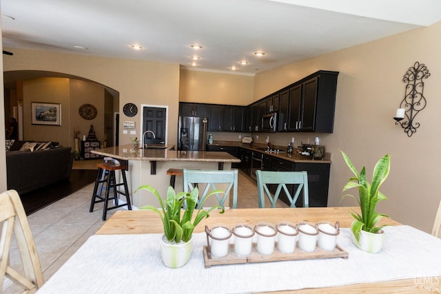 kitchen with a breakfast bar, light tile patterned floors, an island with sink, appliances with stainless steel finishes, and light stone counters