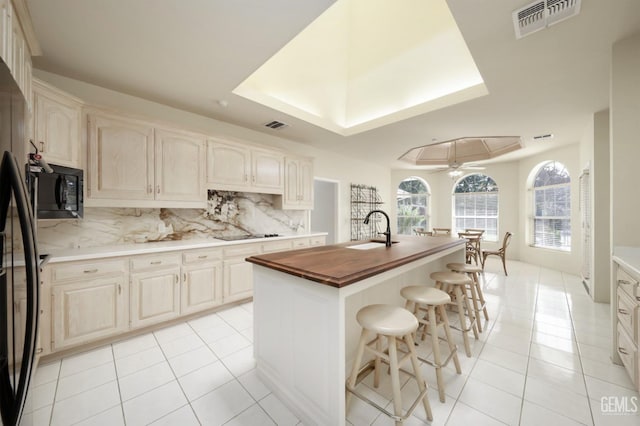 kitchen with light tile patterned flooring, wood counters, tasteful backsplash, a center island with sink, and black appliances