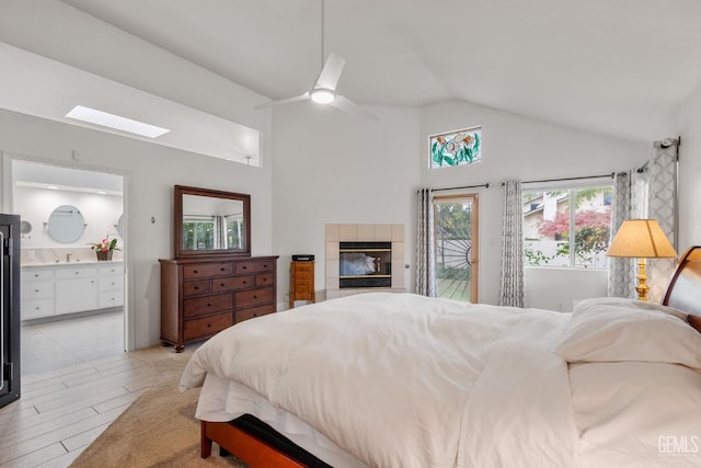 bedroom featuring lofted ceiling with skylight, access to outside, ceiling fan, connected bathroom, and a tiled fireplace