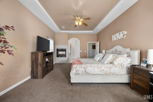 bedroom with dark colored carpet, a multi sided fireplace, ceiling fan, and a tray ceiling