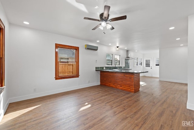 kitchen with hardwood / wood-style floors, an AC wall unit, ceiling fan, wall chimney exhaust hood, and kitchen peninsula