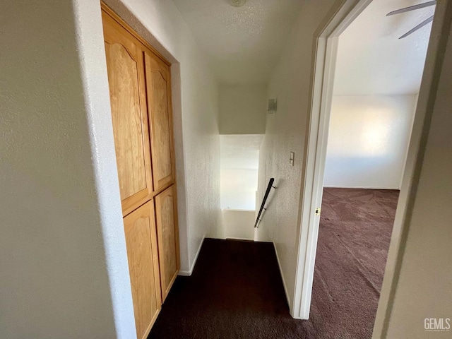 corridor featuring a textured wall, an upstairs landing, baseboards, and dark carpet