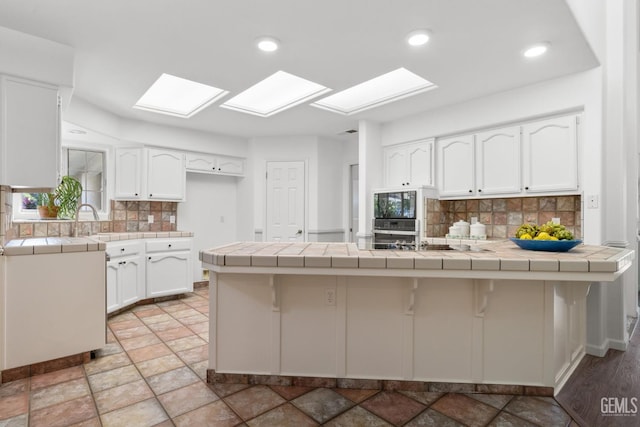 kitchen with decorative backsplash, a peninsula, tile countertops, white cabinetry, and a skylight