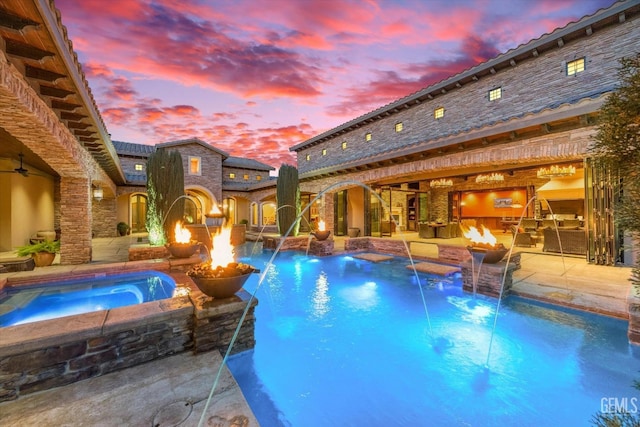 pool at dusk with pool water feature, an outdoor fire pit, and an in ground hot tub