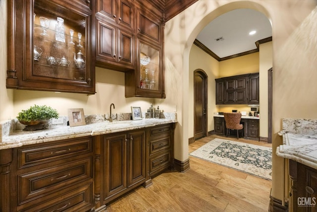 bar with light stone counters, dark brown cabinetry, built in desk, ornamental molding, and light hardwood / wood-style floors