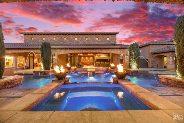 pool at dusk with pool water feature, an outdoor kitchen, a patio area, an in ground hot tub, and a fire pit