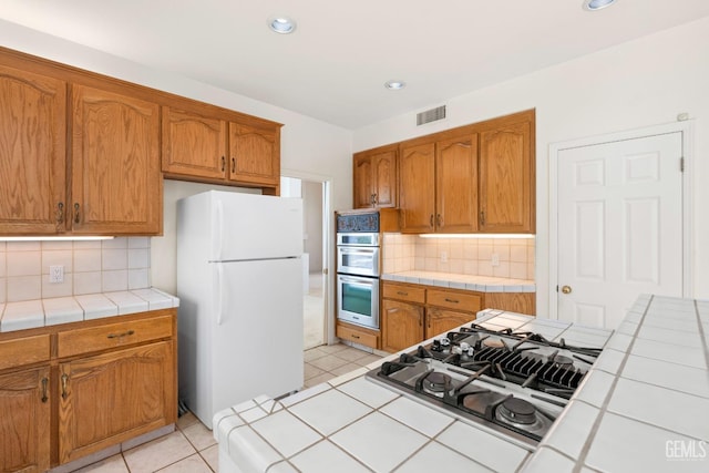 kitchen featuring visible vents, stainless steel appliances, tile countertops, brown cabinetry, and backsplash