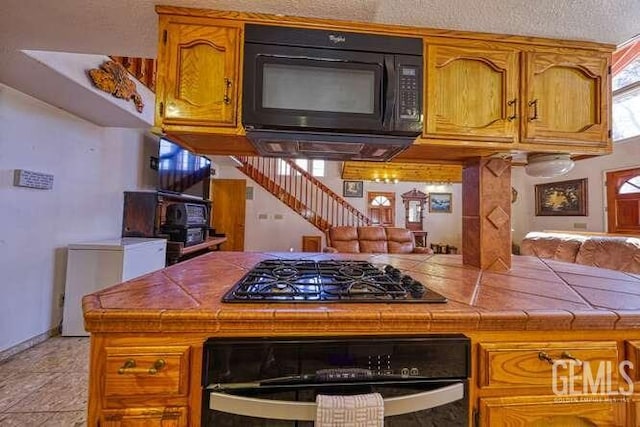 kitchen with wall oven, black microwave, tile counters, brown cabinetry, and gas stovetop