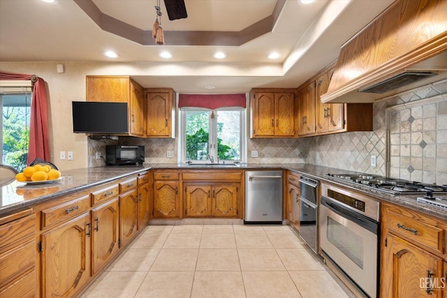 7858 Stine Rd, Bakersfield, CA 93313 - kitchen featuring custom range hood, brown cabinetry, appliances with stainless steel finishes, and a tray ceiling