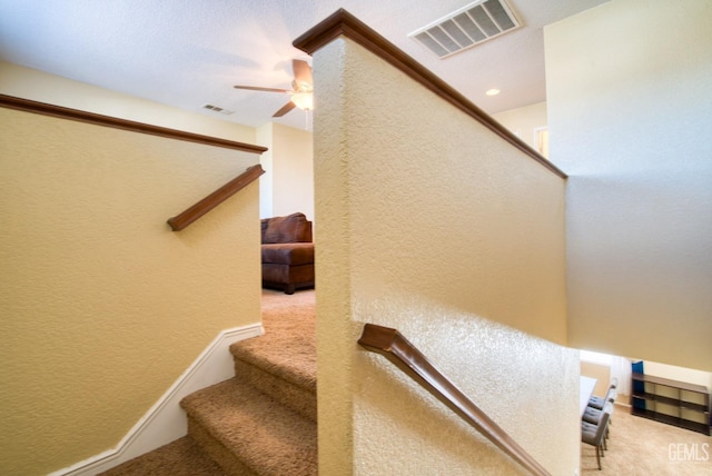 staircase with carpet floors, visible vents, a textured wall, and ceiling fan