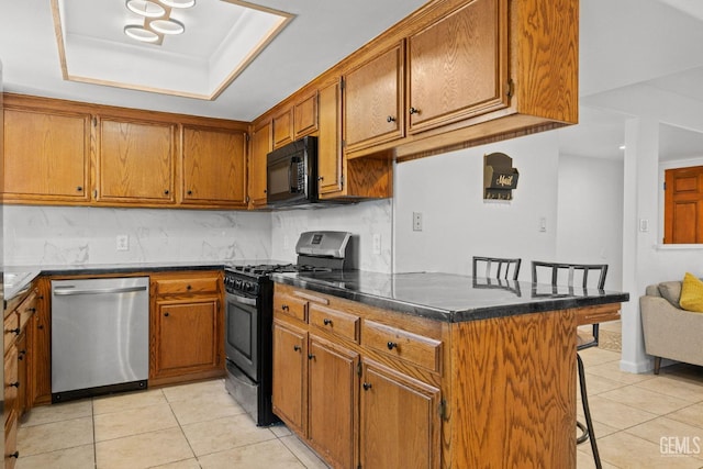 kitchen with dark countertops, appliances with stainless steel finishes, a kitchen breakfast bar, brown cabinetry, and light tile patterned floors