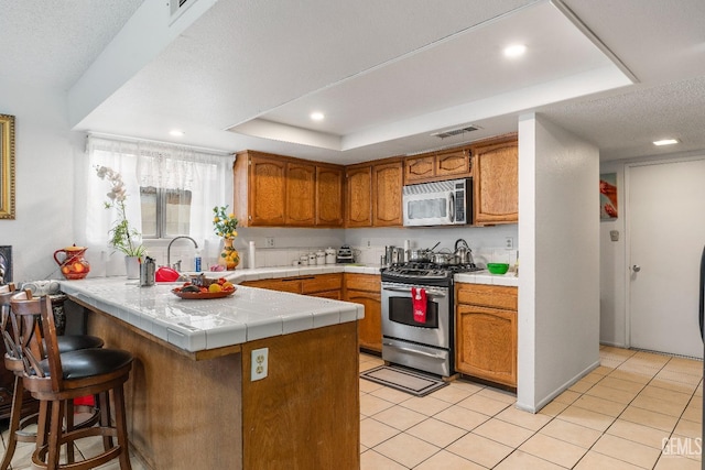 kitchen with a raised ceiling, light tile patterned floors, tile counters, kitchen peninsula, and stainless steel appliances