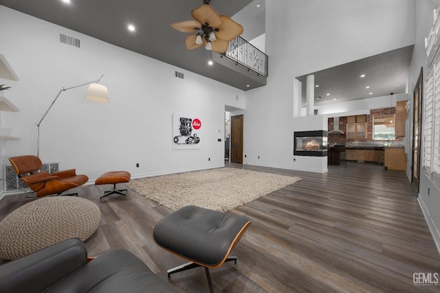 living room featuring dark wood-style flooring, a towering ceiling, ceiling fan, visible vents, and a multi sided fireplace