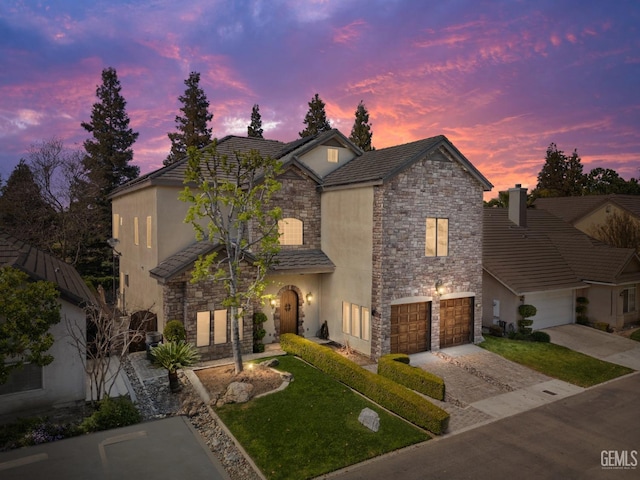 french country style house featuring stone siding, an attached garage, a front yard, stucco siding, and driveway