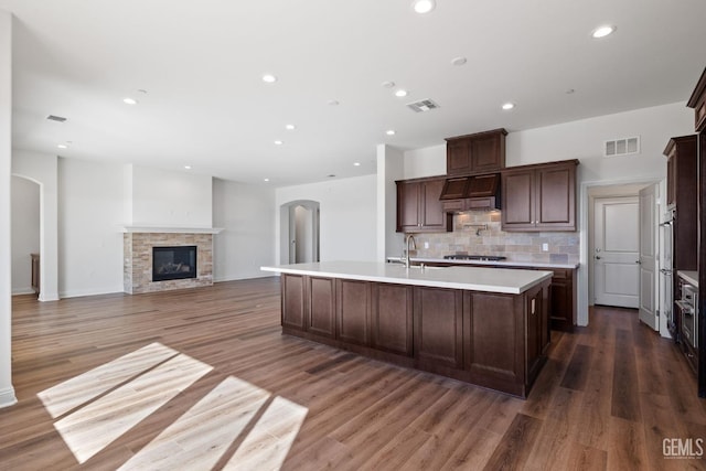 kitchen featuring custom exhaust hood, stainless steel gas stovetop, an island with sink, and dark hardwood / wood-style flooring