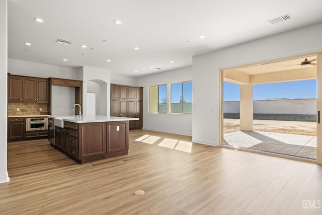 kitchen with tasteful backsplash, an island with sink, sink, stainless steel oven, and light hardwood / wood-style flooring