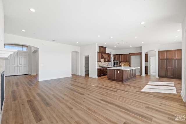 kitchen with decorative backsplash, sink, stainless steel oven, light wood-type flooring, and an island with sink