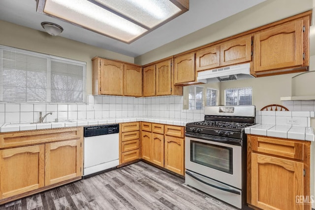 kitchen with tile countertops, stainless steel gas stove, a sink, dishwasher, and under cabinet range hood