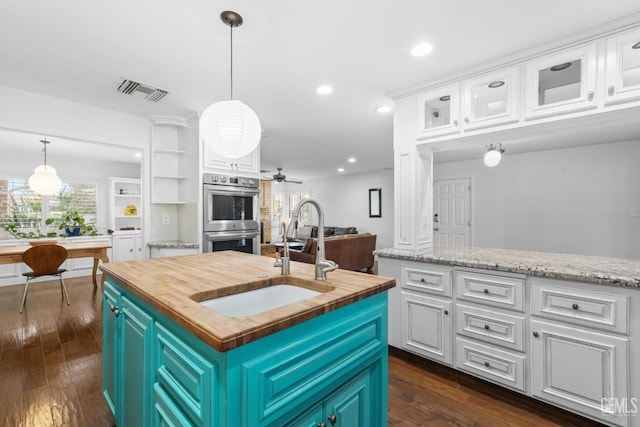 kitchen with an island with sink, butcher block countertops, double oven, white cabinetry, and a sink