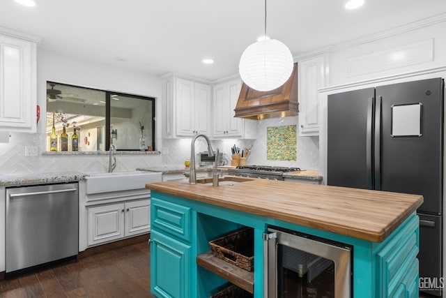 kitchen featuring beverage cooler, an island with sink, butcher block counters, appliances with stainless steel finishes, and white cabinetry