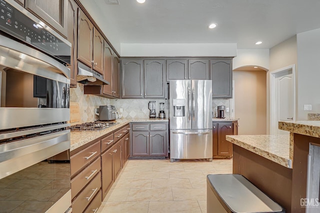 kitchen featuring under cabinet range hood, light stone counters, decorative backsplash, arched walkways, and stainless steel appliances