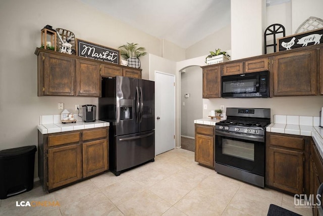 kitchen featuring tile countertops, arched walkways, lofted ceiling, dark brown cabinets, and black appliances