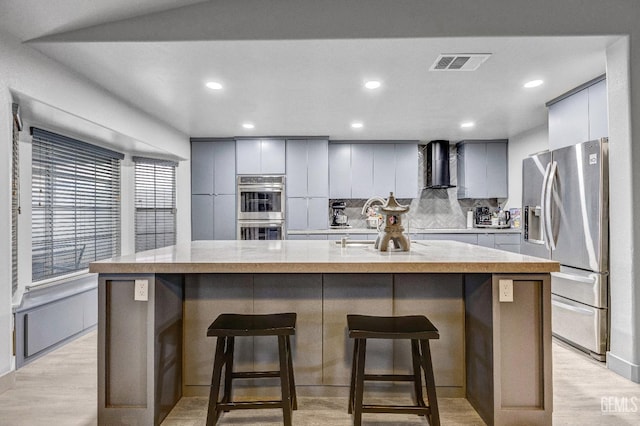 kitchen featuring wall chimney range hood, gray cabinets, a breakfast bar, stainless steel appliances, and an island with sink