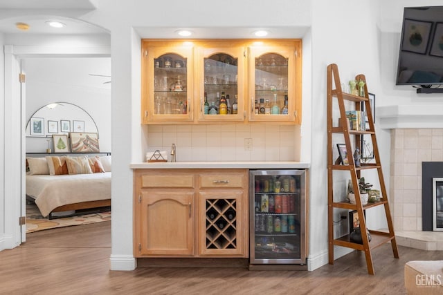 bar with a tile fireplace, light brown cabinetry, decorative backsplash, light wood-type flooring, and wine cooler