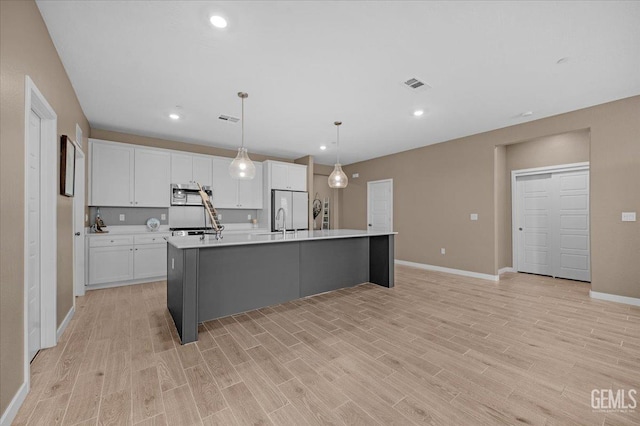 kitchen featuring visible vents, a kitchen island with sink, stainless steel microwave, white cabinetry, and light countertops