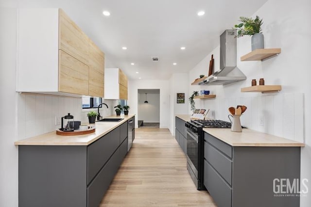 kitchen featuring open shelves, a sink, light wood-style flooring, wall chimney exhaust hood, and appliances with stainless steel finishes