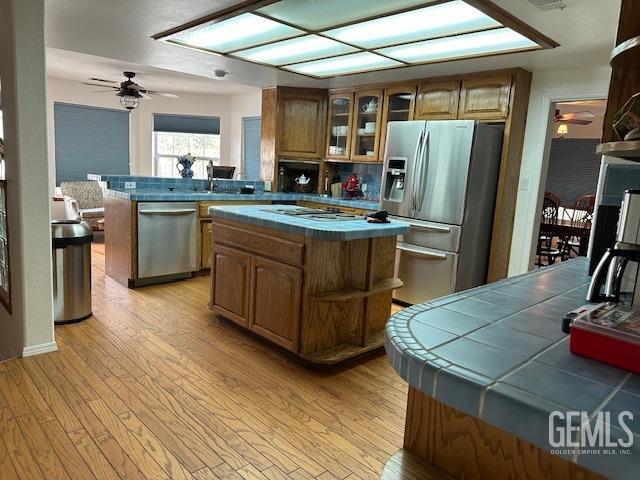 kitchen with ceiling fan, light wood-type flooring, appliances with stainless steel finishes, tile counters, and a kitchen island