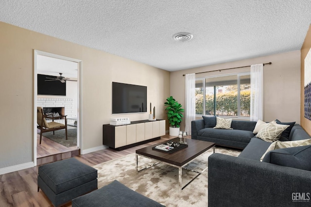 living room featuring a brick fireplace, wood finished floors, a ceiling fan, a textured ceiling, and baseboards