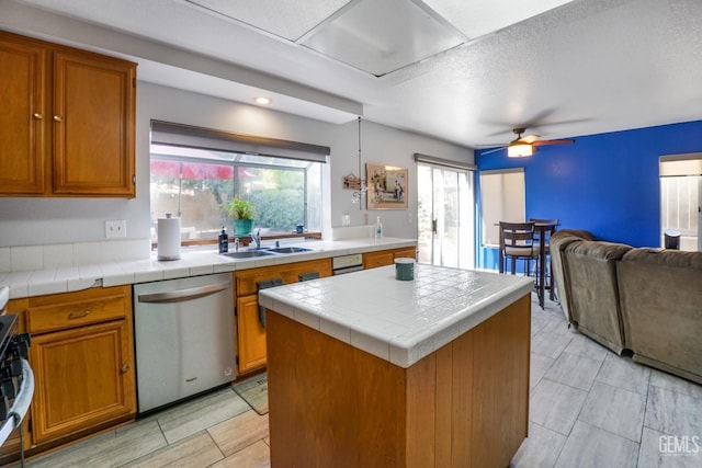 kitchen with tile countertops, a sink, a kitchen island, open floor plan, and stainless steel dishwasher