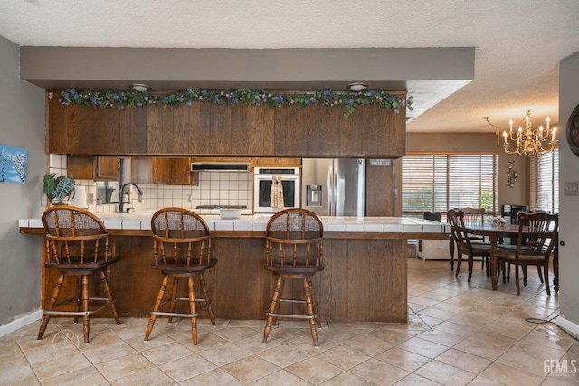 kitchen with wall chimney exhaust hood, a peninsula, a notable chandelier, tile countertops, and appliances with stainless steel finishes