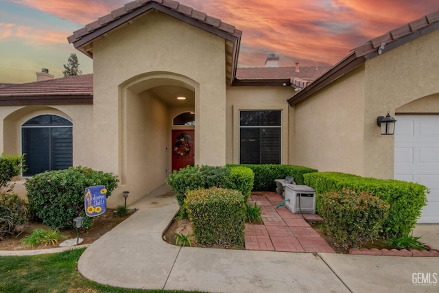 view of exterior entry with a tiled roof, an attached garage, a chimney, and stucco siding