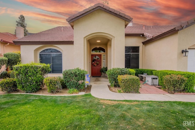 exterior entry at dusk featuring stucco siding, a yard, and a tiled roof