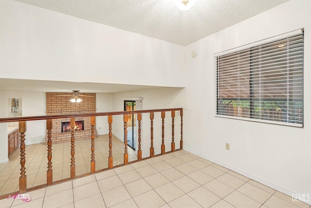 empty room with ceiling fan, a brick fireplace, light tile patterned flooring, and a textured ceiling