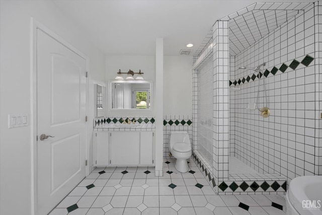 bathroom featuring tile patterned flooring, toilet, tiled shower, vanity, and tile walls