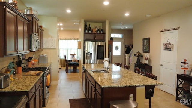 kitchen featuring a center island with sink, a kitchen breakfast bar, appliances with stainless steel finishes, light tile patterned flooring, and light stone counters