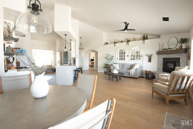 dining area with ceiling fan, light wood finished floors, arched walkways, a brick fireplace, and high vaulted ceiling