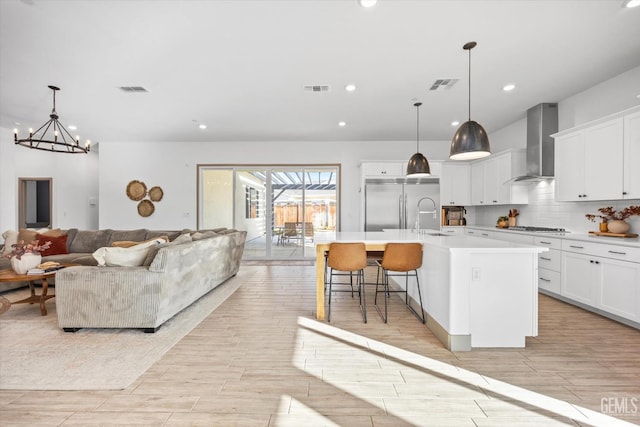 kitchen with hanging light fixtures, white cabinetry, a kitchen island with sink, and wall chimney exhaust hood