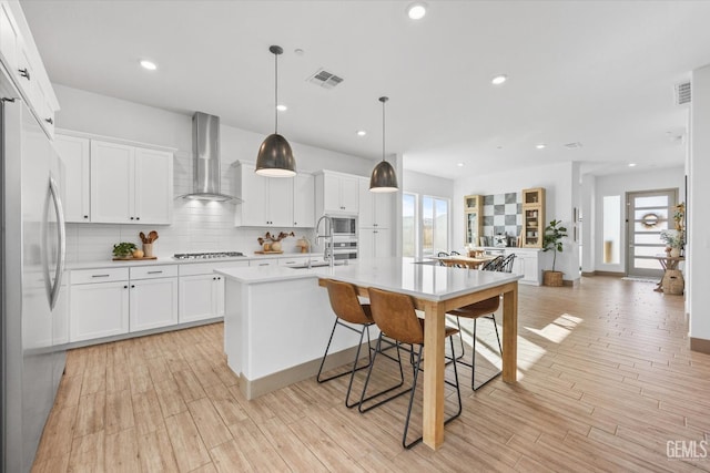 kitchen featuring white cabinetry, wall chimney exhaust hood, an island with sink, pendant lighting, and appliances with stainless steel finishes