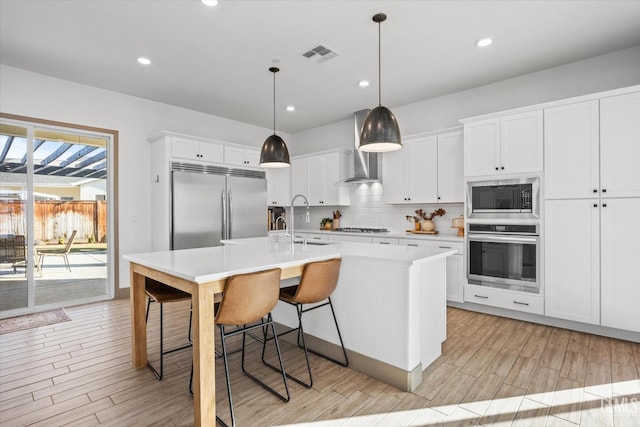 kitchen with wall chimney range hood, pendant lighting, built in appliances, white cabinetry, and an island with sink