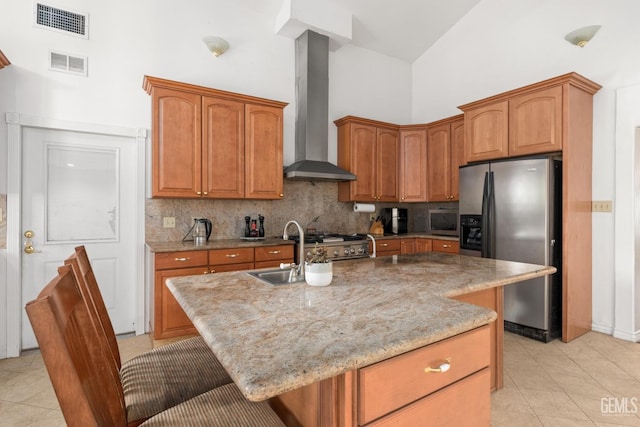 kitchen with wall chimney range hood, sink, stainless steel appliances, light stone countertops, and an island with sink